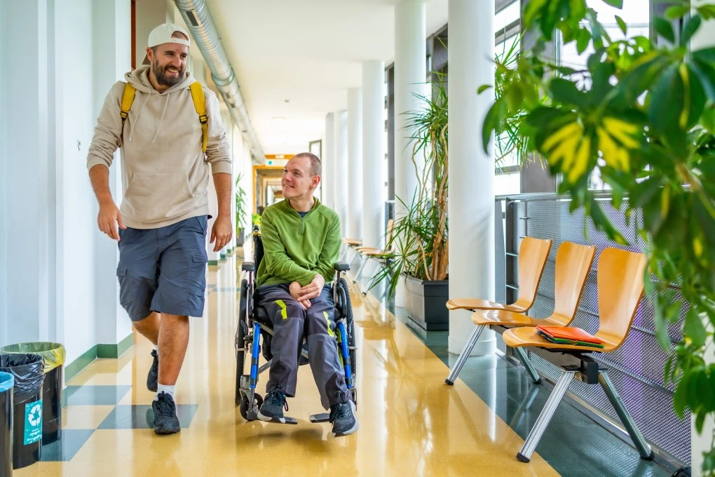 Support worker walking next to disabled man in wheelchair down corridor